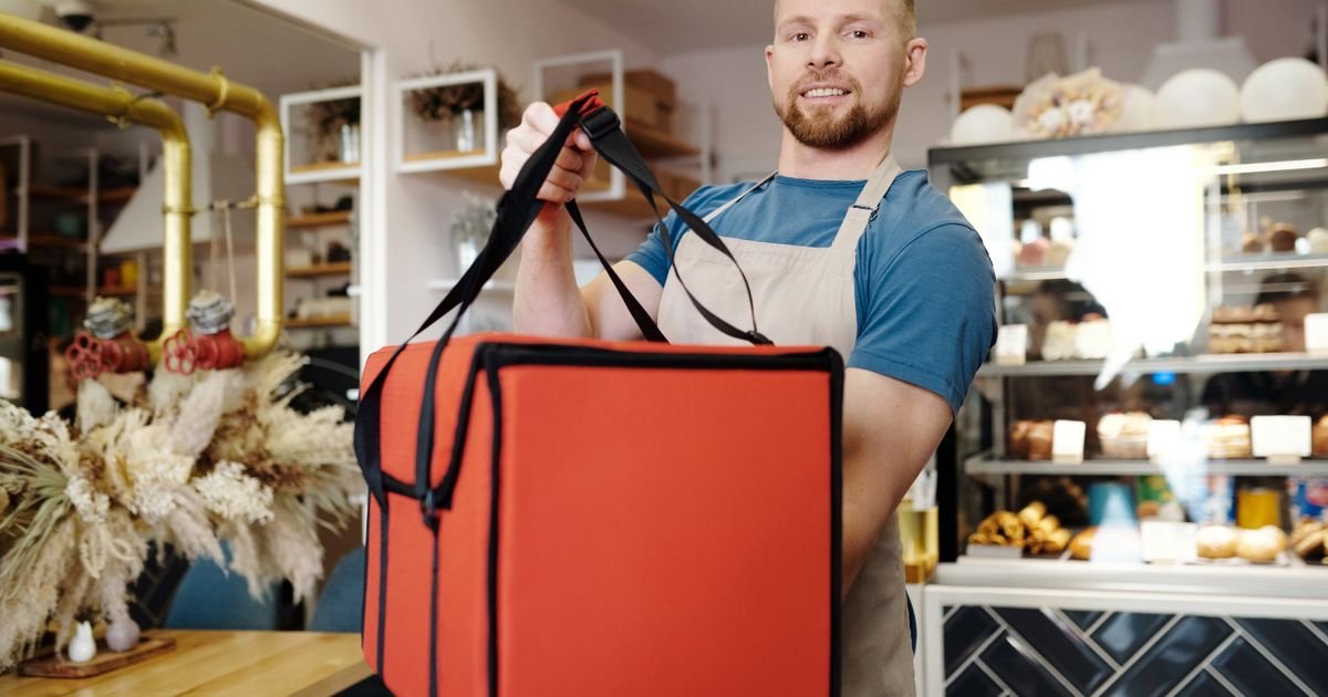 A smiling UK food-business owner in an apron holding a large insulated delivery bag, with a pastry display case behind — Price It Right's guide to selling on Deliveroo, Just Eat and Uber Eats