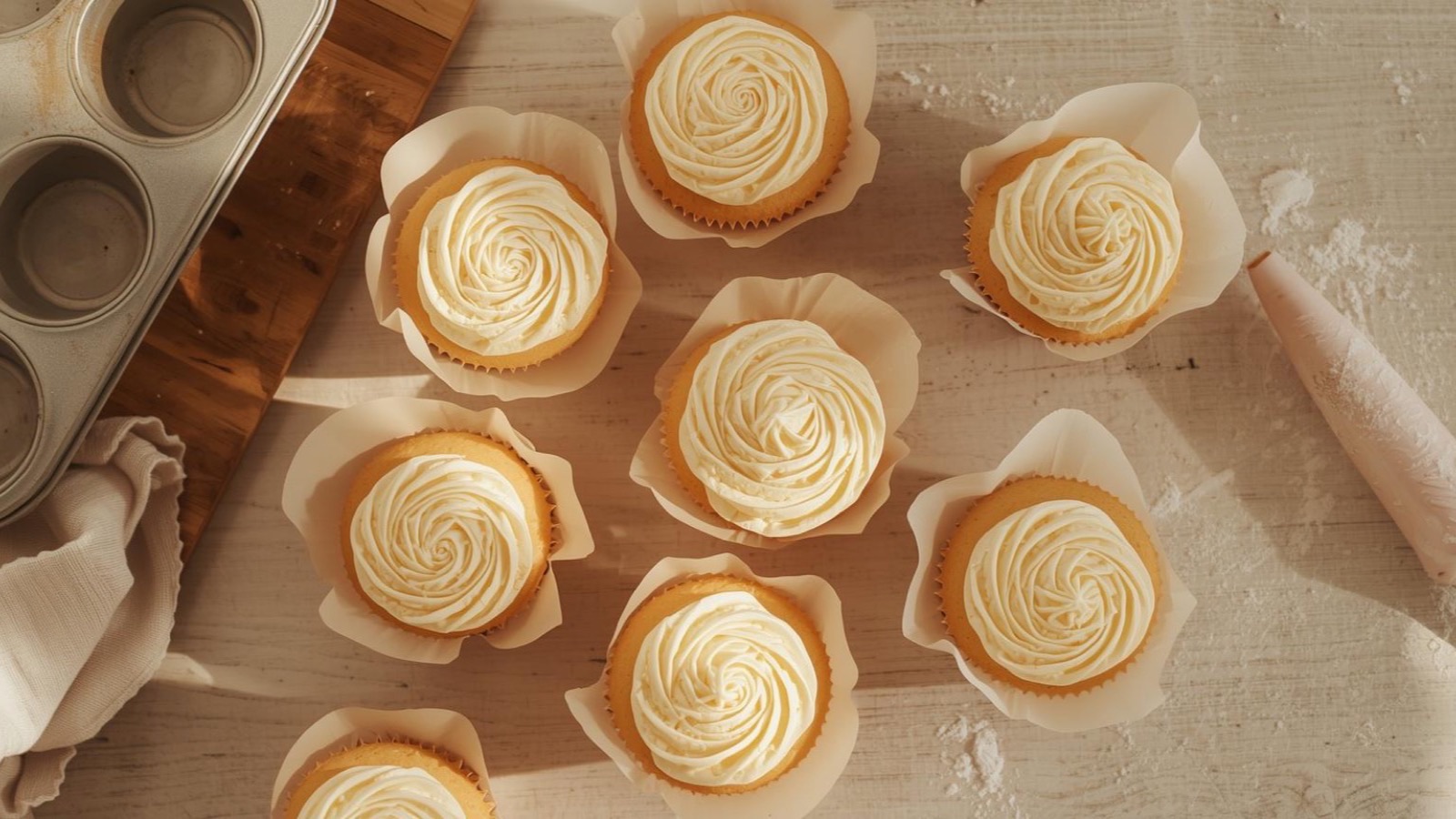 Overhead view of freshly baked home-made vanilla cupcakes with piped buttercream rosettes in white paper liners, shown on a pale wooden surface with a muffin tin and piping bag alongside